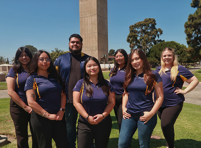 Group of seven people standing outside on a lawn in front of Storke Tower at UCSB